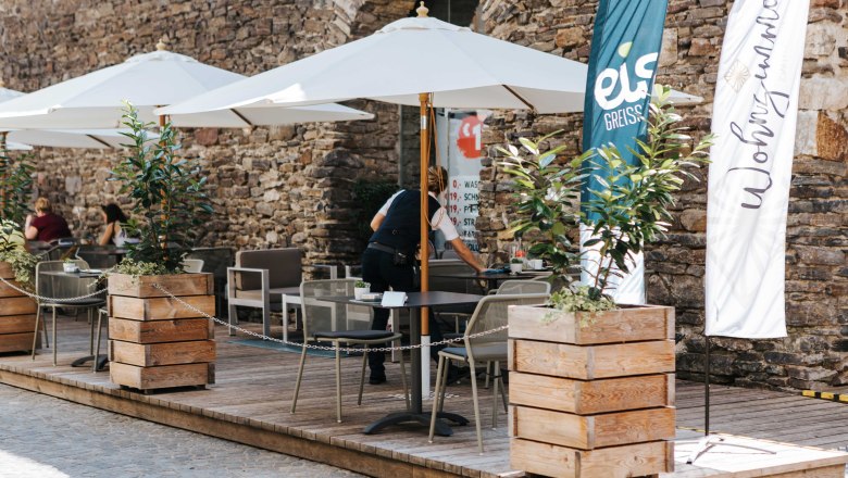 Café living room exterior view, © FMS Steinertor Gastronomie BetriebsgmbH Outdoor area of a café with parasols and wooden crates as planters in front of a stone wall.