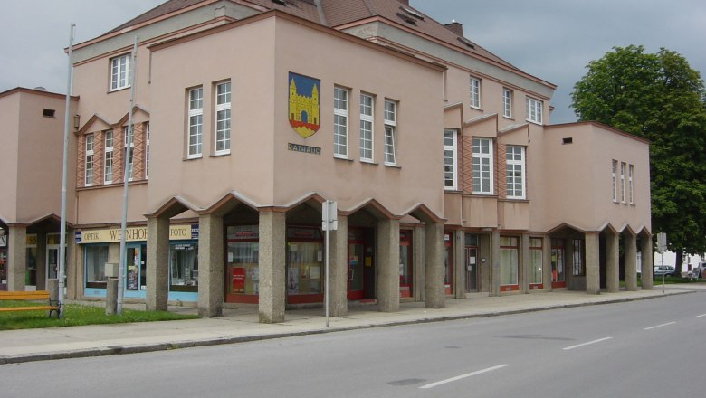 Town hall, © Gemeinde Hohenau/ March A modern town hall building with a pink façade and large windows. It has a coat of arms on the corner and stores on the first floor.