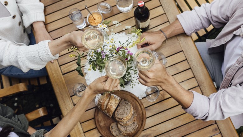 Heurigen snack, © Weinviertel Tourismus GmbH / Sophie Menegaldo Four people clink glasses of wine at a wooden table with bread and flowers on it.