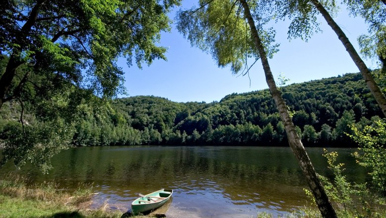 Kampsee Dobra, © Best Trip A calm lake with a rowing boat on the shore, surrounded by densely wooded hills and clear skies.