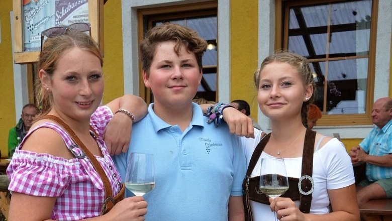 Schabasser family, © Schabasser Three young people in traditional Bavarian dress with glasses of wine in front of a yellow building.