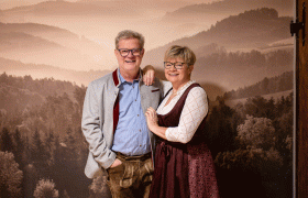 Petra and Alfred Weber, © Thomas Gobauer - VitalZeit Hotel Weber A couple in traditional Bavarian dress stands in front of a background with a forest landscape.