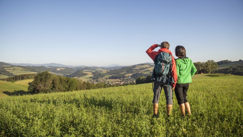 Along the path of glances, © Gemeinde Bad Schönau, Stefan Knittl Two people are standing in a meadow, gazing into the distance over a hilly landscape.