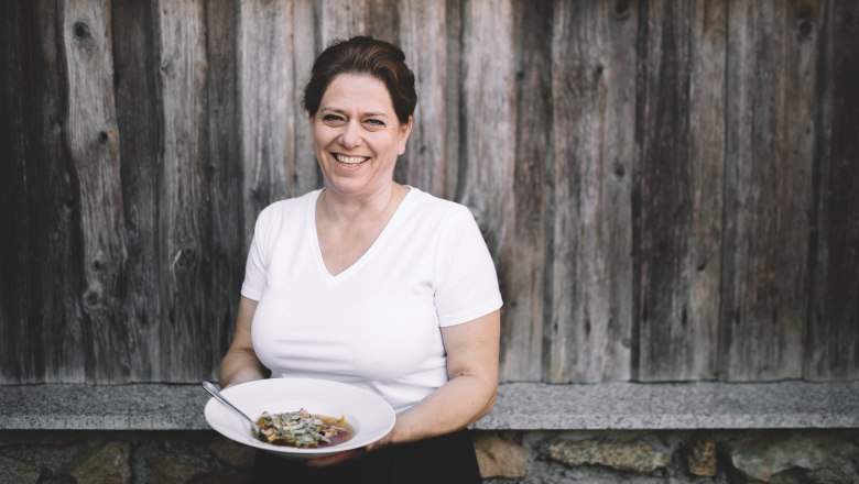 Landlady Doris Schreiber, © Niederösterreich Werbung/Mara Hohla Woman in white T-shirt holding a plate of food in front of a wooden wall.