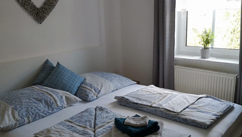 Room 1, © Haselberger A cozy bedroom with a double bed, decorated with blue bed linen and towels. A picture of a heart hangs on the wall, a window with a curtain next to it.