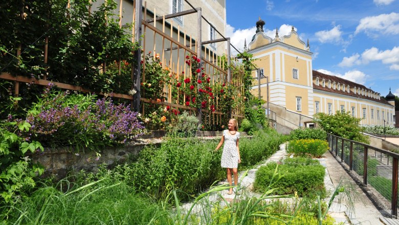 Zwettl Abbey Gardens, © Waldviertel Tourismus, Robert Herbst A woman in a white dress stands in a flowering garden in front of a historic building with a yellow façade and towers.