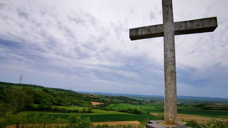 Cone cross, © Weinstraße Weinviertel A large stone cross on a hill overlooking a green landscape and cloudy sky.