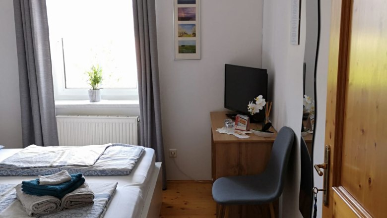 Room 1, © Haselberger Bright bedroom with double bed, gray curtains, small desk and chair, TV and carpet on wooden floor.