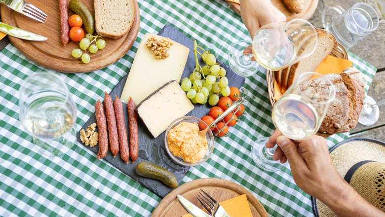 Heurigen snack, © Weinviertel Tourismus GmbH / Schwarz-König Picnic table with cheese, bread, sausage, grapes and wine on a checkered tablecloth.