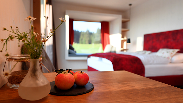 roon_room2, © Franz Atteneder A modern hotel room with a red bedspread, a table with apples and a window with a view of the greenery.