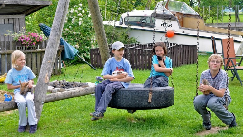 Children in the garden, © Kerndlerhof Four children sit on a swing in the garden and hold animals. There is a boat in the background.