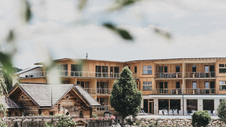 Naturhotel Molzbachhof, © Heldentheater, Hotel Molzbachhof A modern wooden building with balconies and a traditional wooden house in the foreground, surrounded by trees and gardens.