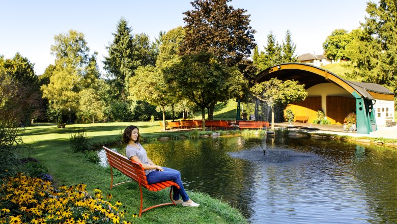 Bad Schönau spa gardens, © Niederösterreich Werbung/Doris Schwarz-König Woman sitting on a red bench by the pond in the spa gardens of Bad Schönau with trees and pavilion in the background.