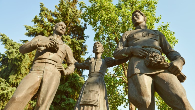 Peace Monument, © Klaus Engelmayer Three bronze statues, two men and a woman, stand next to each other in front of trees.
