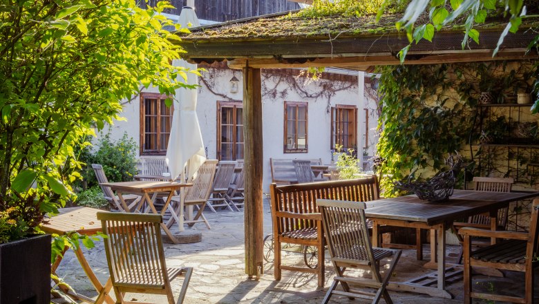 Bed in the Wies'n, © Niederösterreich Werbung / Maximilian Pawlikowsky A sunny garden with wooden furniture and a covered seating area, surrounded by plants and a building in the background.