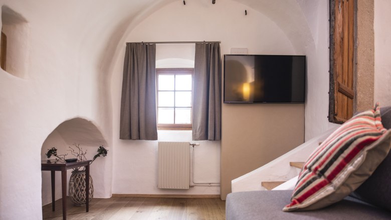 Residence Ap. 3, © Daniela Matejschek Interior view of a cozy room with vaulted ceiling, window with curtains, flat screen TV and sofa with striped cushions.