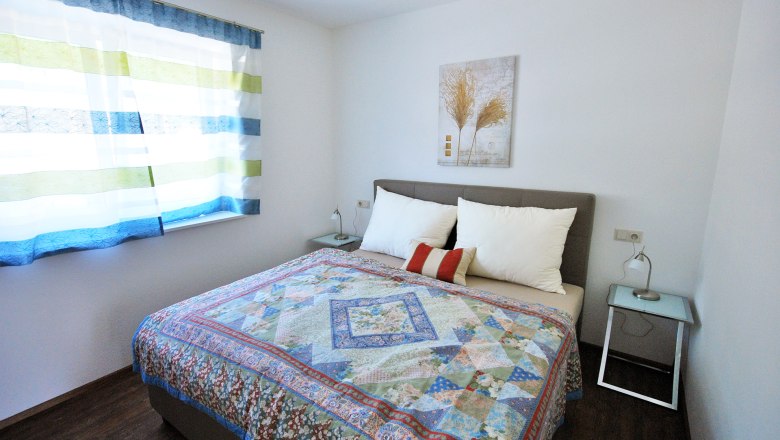 Bedroom 2, © Familie Harrauer A bedroom with a double bed, colorful bedspread, two bedside tables and a window with striped curtains.