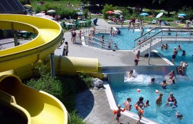 Parkbad Haag, © zVg Gemeinde Haag An outdoor pool with a yellow water slide, swimmers in the pool and people on the sunbathing lawn.