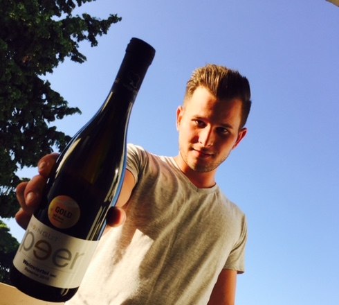 Patrick Peer, © Weingut Peer A man holds a bottle of wine up to the camera, blue sky and trees in the background.