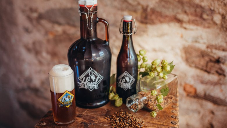 Hazel brew, © Niederösterreich Werbung/Daniela Führer Beer bottles and glass with Haselbräu logo, hops and malt on wooden table.