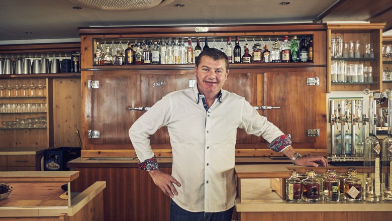 Landlord Markus Hawlik, © Niederösterreich Werbung/Andreas Hofer A man in a white shirt stands behind a bar with bottles and glasses in the background.