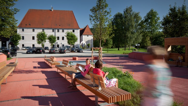 Municipality of Zeillern, © Kurt Hörbst A modern square with wooden benches and people in a sunny setting, with a large building with a red roof in the background.