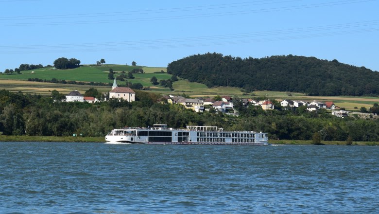 Exterior view2, © Gasthof Donaublick A river cruise ship sails on a river in front of a green hilly landscape with a village in the background.