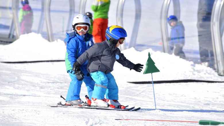 Family ski area, © ©familienarena Two children in ski equipment skiing in the snow, accompanied by an adult in the background.