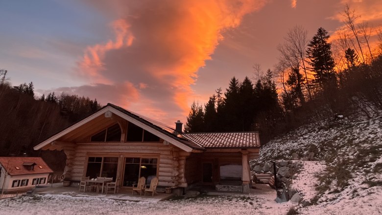 RaxChalet in winter, © RaxChalet A wooden chalet in the snow at sunset with an orange sky and trees in the background.