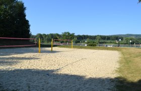 TD beach volleyball court, © Stadtgemeinde Korneuburg An empty beach volleyball court with yellow posts and a pink net, surrounded by trees and a river in the background.