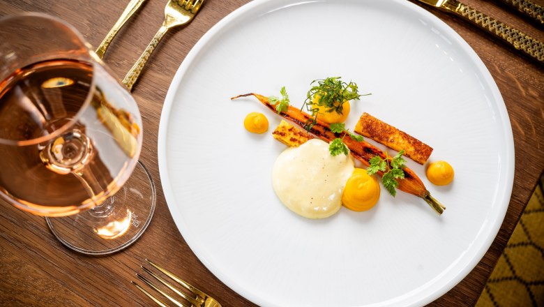 Carrot-polenta-hay-chervil-wine, © Knappenhof Christian Husar An elegant dish of carrots, polenta, hay and chervil on a white plate, next to a glass of rosé wine on a wooden table.