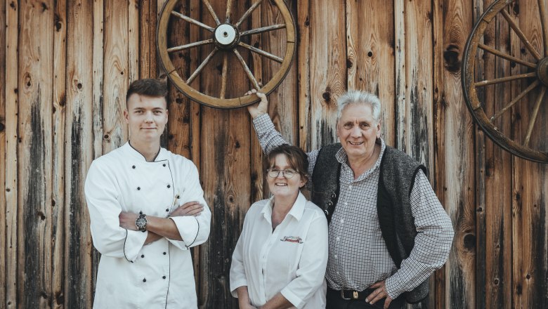 Mayrhofer host family, © Niederösterreich Werbung/David Schreiber Three people in front of a wooden wall with wagon wheels, one person in cooking clothes.