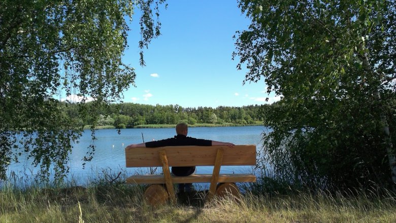 Langau mining lake, © Hannes Messmann Person sitting on a bench on the shore of a lake, surrounded by trees.