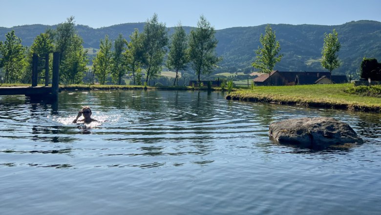 Swimming, © Familie Moser Person swimming in a pond with trees and hills in the background.