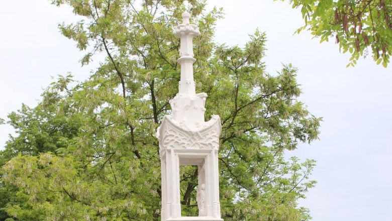 Guntersdorf, © Gemeinde Guntersdorf White stone column with cross in front of a green tree.