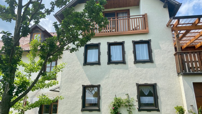 Apartments Riedl, © Wiener Alpen A traditional house with white walls, wooden cladding and a balcony, surrounded by green trees and plants.