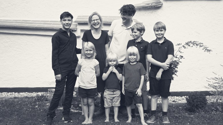 Pölzer family, © Monika Pölzer A family of seven stands smiling in front of a white wall outside.