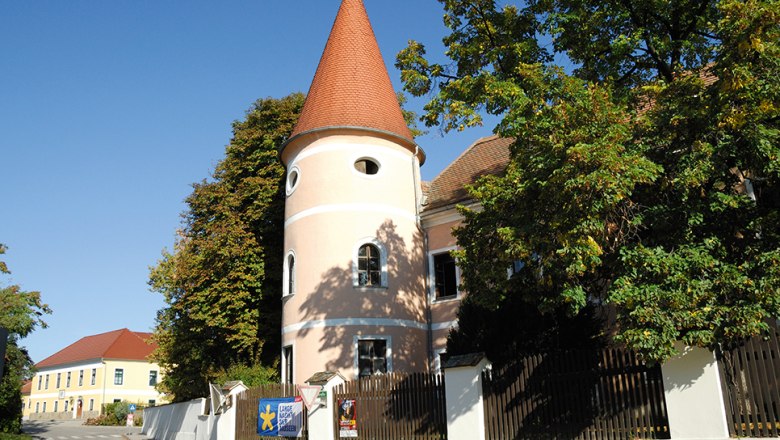 View of Fels am Wagram, © Gemeinde Fels am Wagram A pink building with a tower and red roof in Fels am Wagram, surrounded by trees and a blue sky.