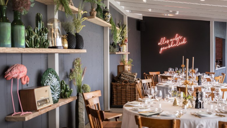 Distant view, © Matthias Kronfuss A stylishly decorated room with laid tables, plants and neon lettering on the wall.