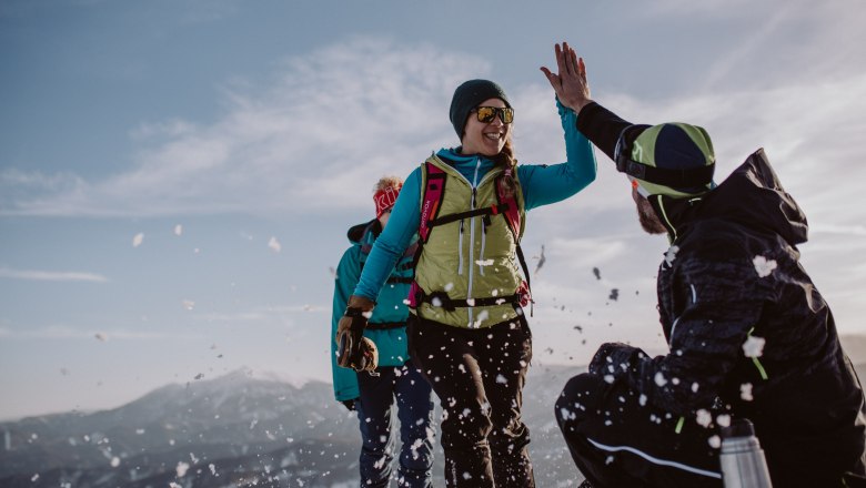 Unterberg ski area, © Schigebiet Unterberg | katischweiger.fotography Three people in the snow, one high-fiving another, with mountains in the background.