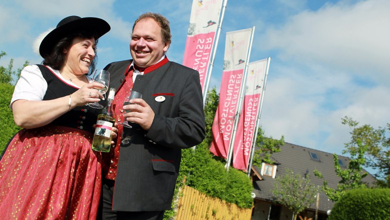 Tavern Moderbacher, © weinfranz.at A man and a woman in traditional dress toast with glasses of wine, flags and a building can be seen in the background.