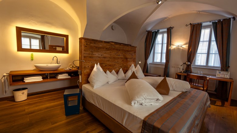 Hotel Landhaus Moserhof, © Niederösterreich Werbung / Maximilian Pawlikowsky Comfortable hotel room with double bed, wooden wall, washbasin and desk in front of two windows.