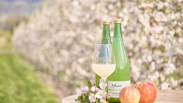 Apple juice, © Philipp Monihart Two bottles of apple juice, a glass, two apples and apple blossoms on a table in front of blossoming apple trees.