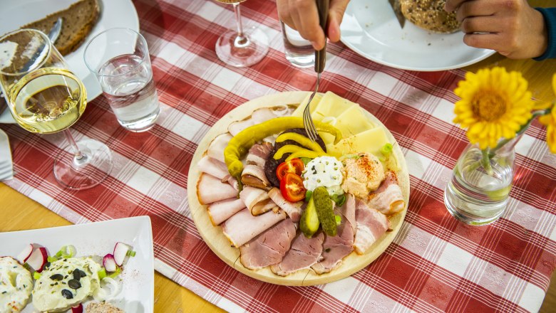Heurigen snack, © POV / Robert Herbst A table set with a platter of cold cuts, bread, wine and flowers on a red and white checkered tablecloth.