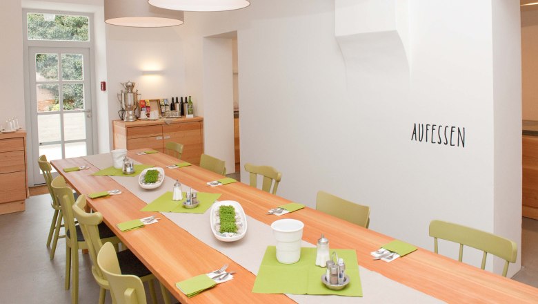 ABOVE, © Stefanie J. Steindl A bright dining room with a long wooden table, green chairs and decorations. 'AUFESSEN' is written on the wall.