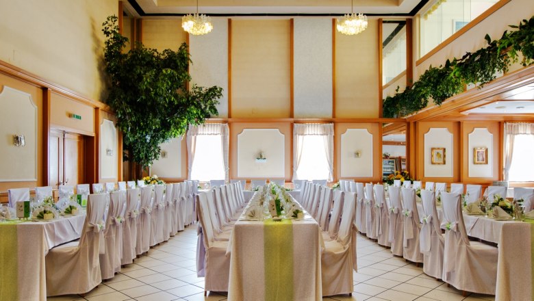 Restaurant, © Auland-Hotel Siebenbrunnerhof Elegant banquet hall with laid tables and white chairs, decorated with plants and chandeliers.