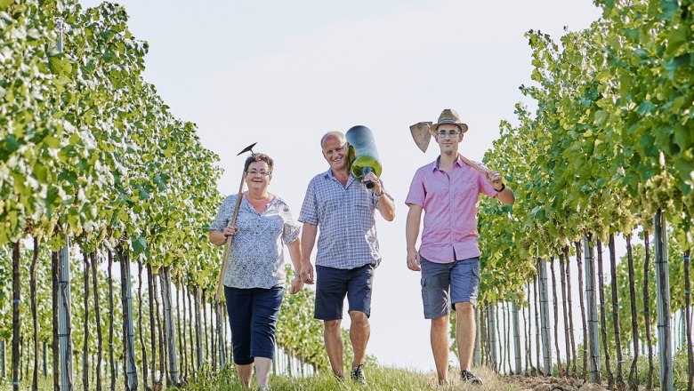 Winegrower family Humer, © Martin Sommer Three people walk through a vineyard, carrying tools and smiling.