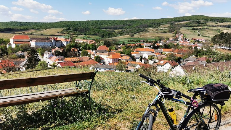 Cycle path to the Hundschupfen vineyard, © Weinstraße Weinviertel Bicycle and bench with a view of a village in a hilly landscape.