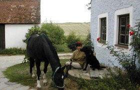 Horse farm Fam. Vasicek, © Vasicek A person sits next to a black dog in front of a blue house while a black horse eats grass.