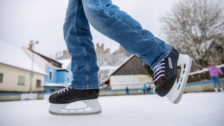Kirchschlag ice rink, © Wiener Alpen, Martin Fülöp Close-up of ice skates on an ice rink with buildings and castle ruins in the background.